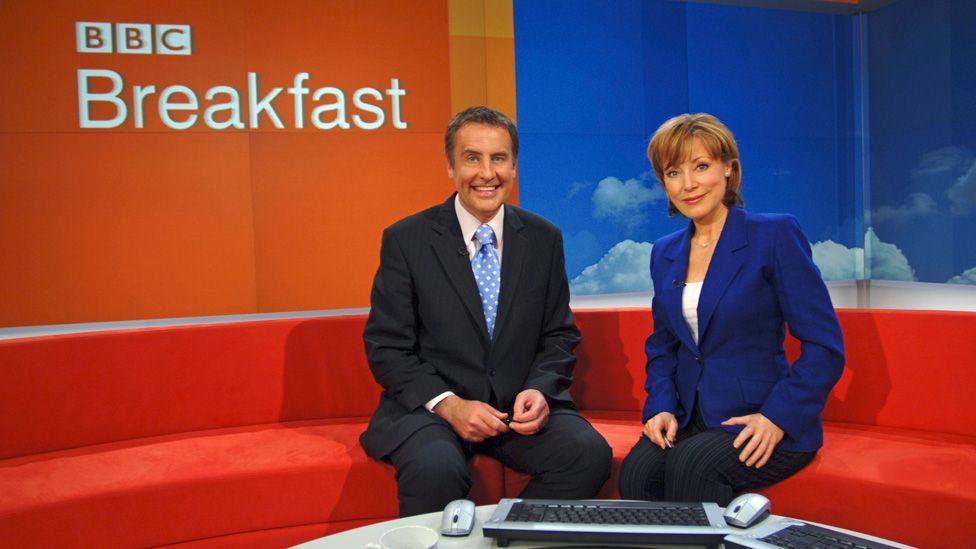 Dermot Murnaghan and Sian Williams posing on the BBC Breakfast red sofa in 2006, in front of a large BBC Breakfast logo on a screen