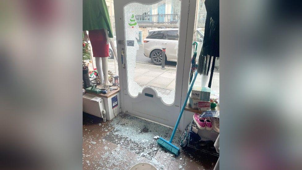 A charity shop from the inside looking at a smashed glass door. The broken glass is on the floor of the shop.