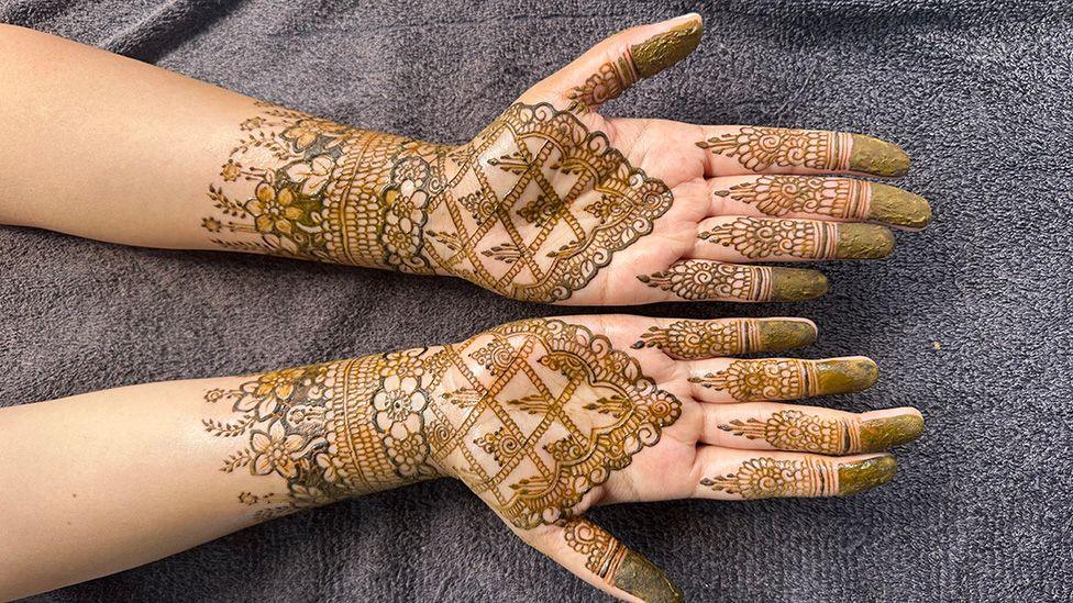 Two upturned hands resting on a grey towel with elaborate floral designs in Henna