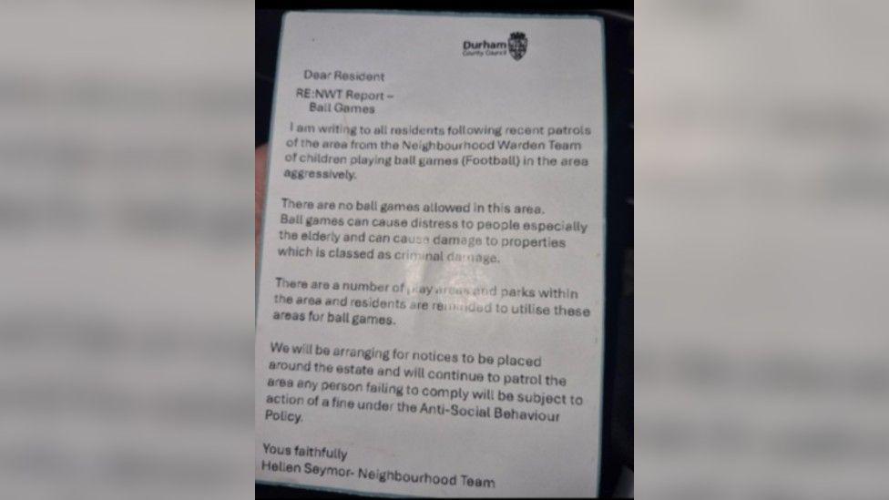 A hand holding a laminated fake note, claiming to be from Durham County Council Neighbourhood Wardens - North team. The letter states it was sent over children playing football aggressively and claims no ball games are allowed in the area. It adds ball games can cause damage to property and distress. It ends with a claim that more patrols will be carried out and notices will be placed in the area. It says those failing to comply will be fined. Durham County Council's logo is printed in the top right corner.