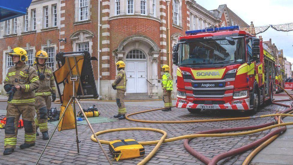 Red and yellow hoses from fire engines lay across a street with firefighters standing either side of a A-frame board.