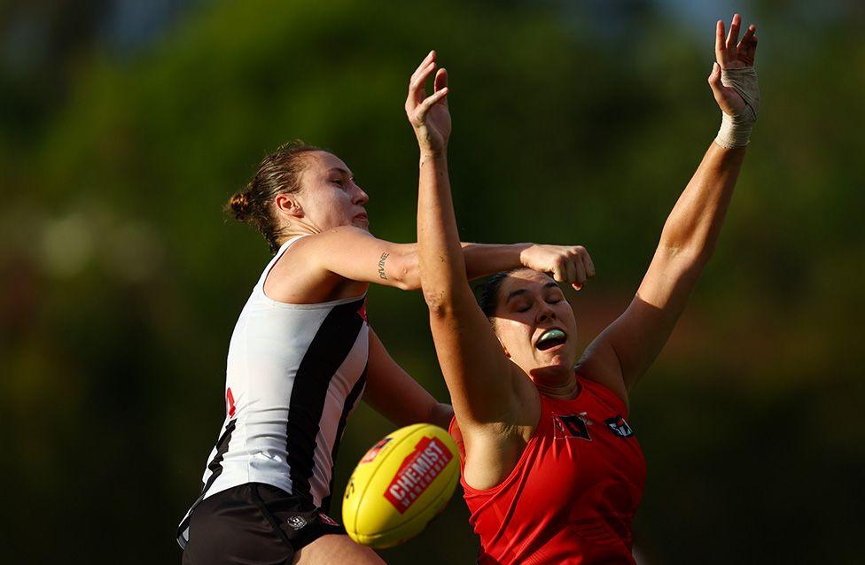 Two female players compete for possession of a yellow Australian rules football during a match. One player in a black and white jersey reaches forward nearly hitting the other player, while the other in a red jersey raises both arms. The background shows green trees under bright sunlight.