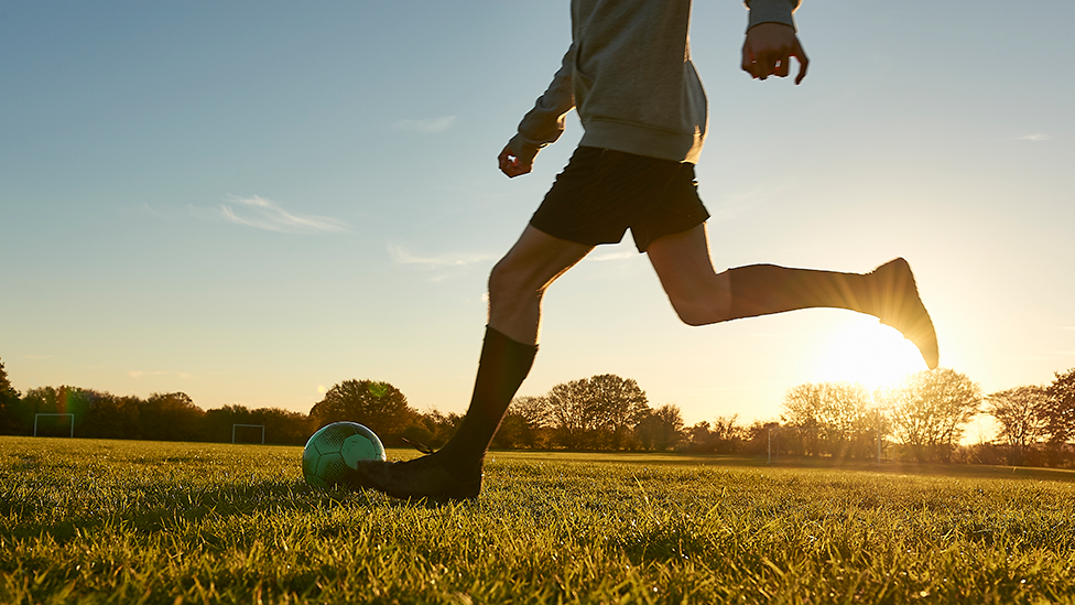 Generic image of a footballer from the waist down dribbing a ball on a field. they are wearing dark boots and socks and dark shorts, with a light grey top
