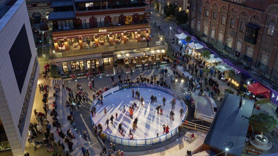 Round ice ring in middle of square with stalls and shops around the edge - dozens of people are milling around and skaters are on the rink.