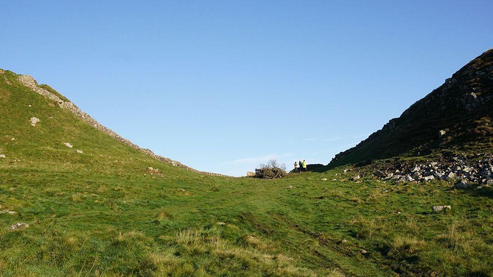 A traditional view of the Sycamore Gap tree, with the coarse, grassy land rising away from the dip, so left and right - except the gap is no longer filled with the tree. Where the tree stood there are just the remaining branches with two people in yellow hi-viz clothing standing nearby.
