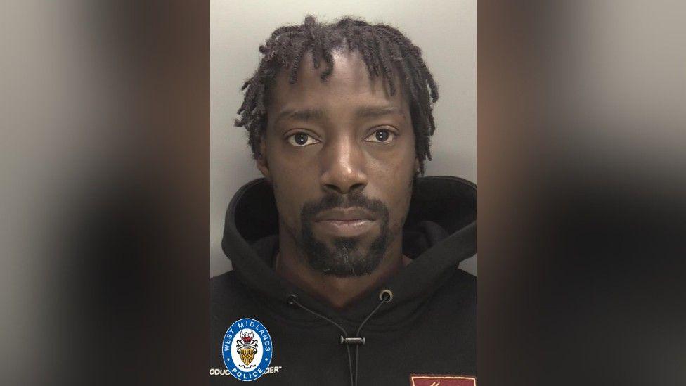 A police mugshot of a man with dark dreadlocks and a black goatee-style beard. He is wearing a black hoodie. A West Midlands Police logo has been superimposed in the corner of the picture.