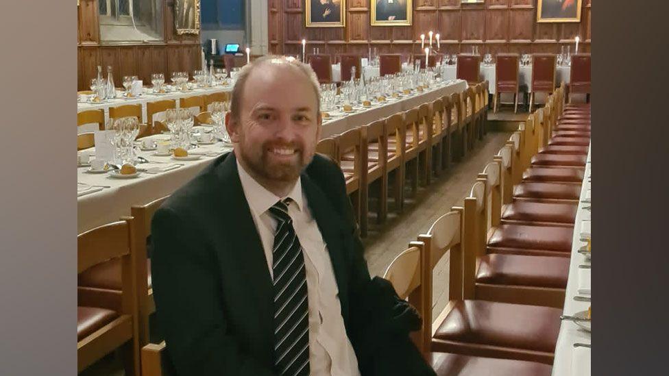 Chris Whitaker, with short fair hair and a close cropped beard, wearing a dark suit, white shirt and striped tie, smiling at the camera, sitting in at a long white clothed dining room, with two more long tables set for a meal behind him in a panelled room