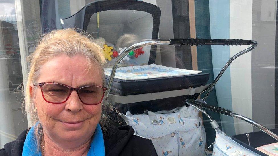 Rachel Green standing in front of a pram outside the Fenland Family and Baby Bank. She has blonde hair, red framed glasses, a black top and a blue scarf.