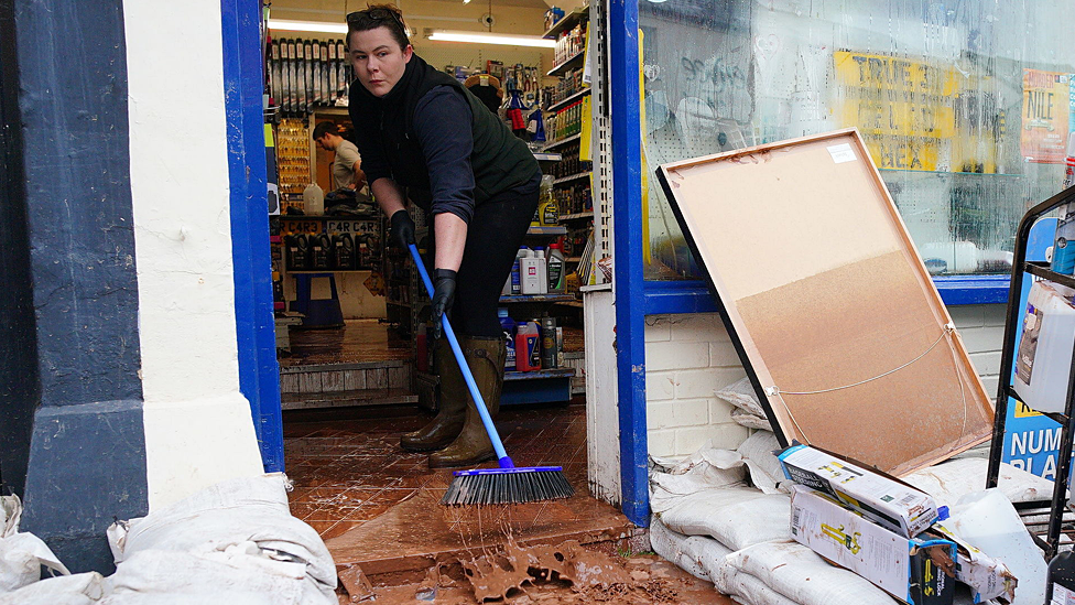 Woman sweeps brown muddy water out of a shop door with a gap made in sandbags over the front step