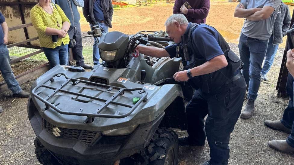 A man in a blue shirt puts his arm onto a grey-coloured quad bike. Behind him a group of about six people can be seen standing around watching