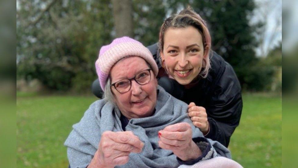 Shareen Elnagy wearing a black jumper with her mum, who us wearing a pink hat and glasses and a grey shawl. There are trees and grass behind them. 