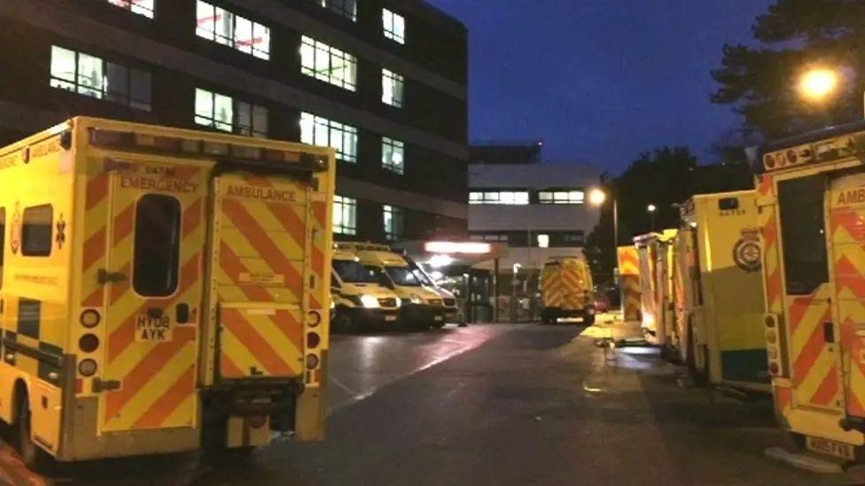 About a dozen ambulances waiting outside the Queen Alexandra hospital in Portsmouth. It is night time and all the lights are on in the hospital.