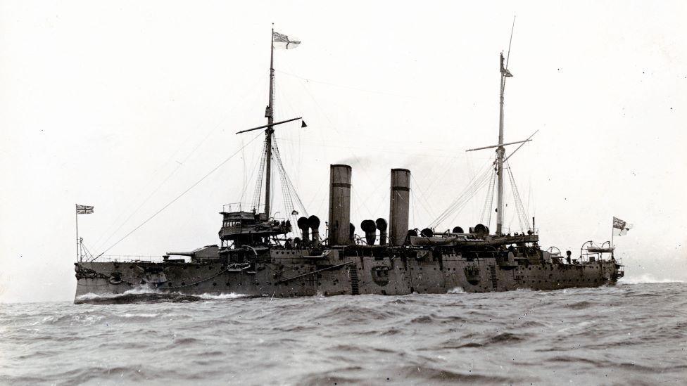 Black and white image of HMS Hawke, a Royal Navy warship, at sea, two funnels and two tall masts, and flags at either end. 