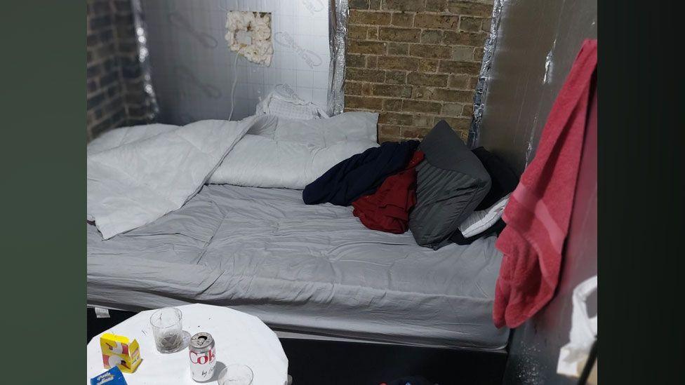 A bed with a light grey sheet, a dark grey pillow and a bundle of clothes, with a red towel hanging on a makeshift wall made of silver insulating panels to its right and a white circular coffee table in front 