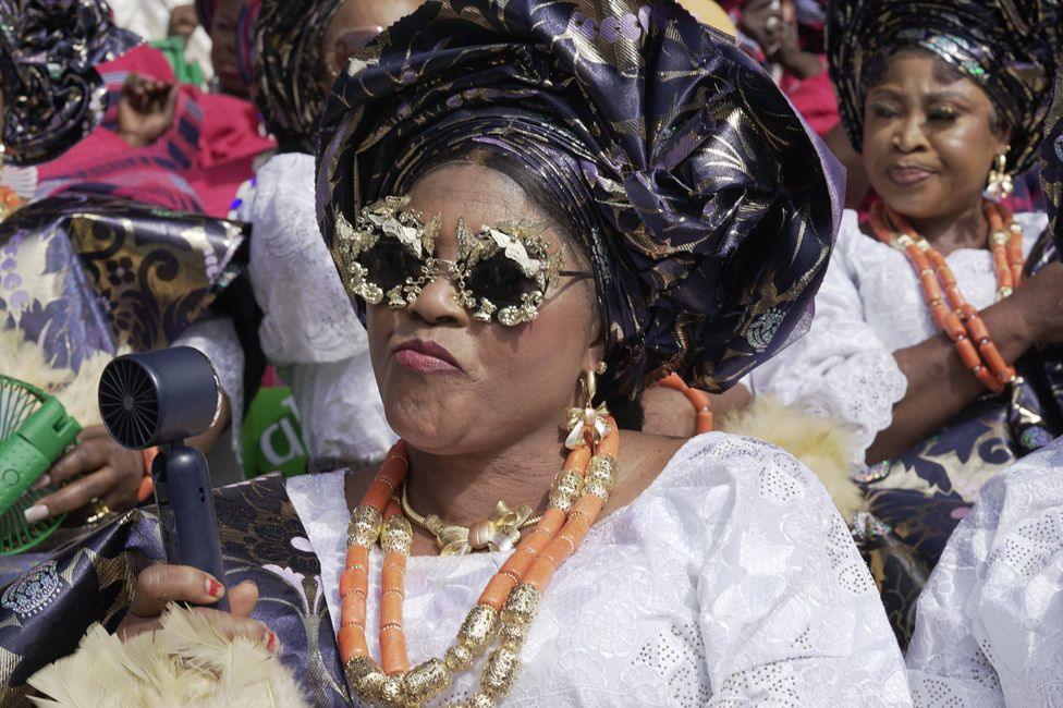 A woman in a dark head wrap and sunglasses holds an electric fan to her face.
