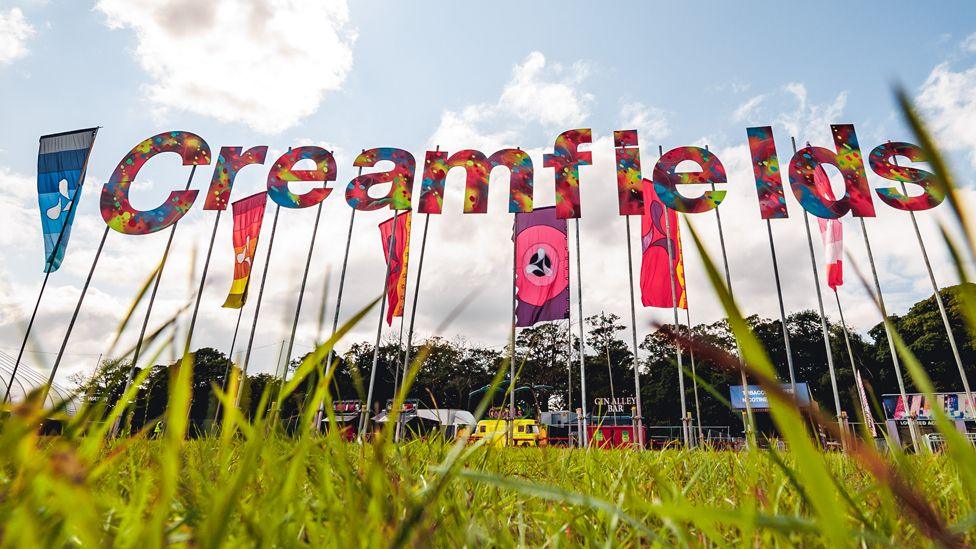 A multi-coloured Creamfields sign stands on poles amongst blue, purple and red flags, slightly obscured by the grass of the festival's site in the foreground