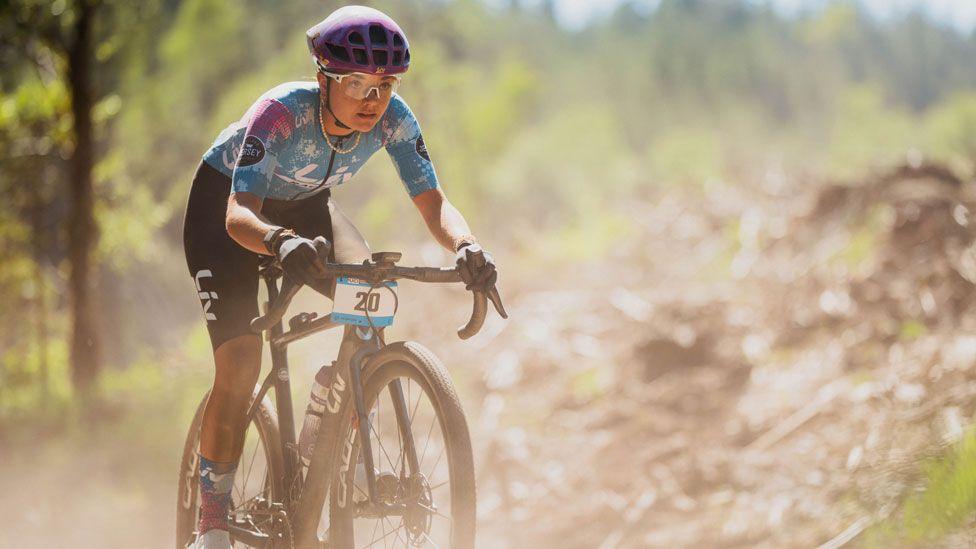 A gravel road racer in a blue and pink top and black shorts speeds through a dusty forest track