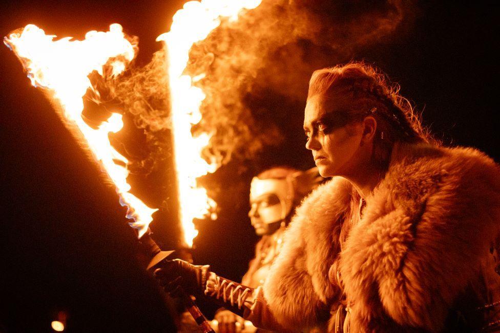 A woman in Celtic inspired dress holding flaming torches. 