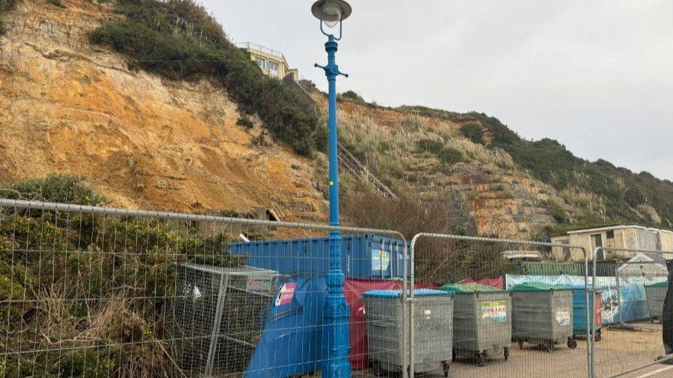 Metal fencing with large bins behind, blue shipping container and cliffs.