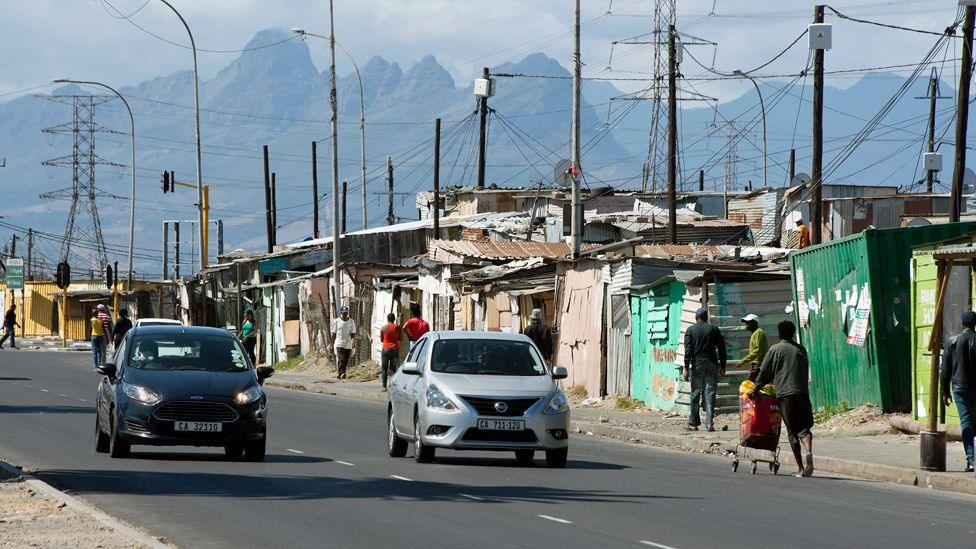 Two cars drive down a main tarmac road in Khayelitsha. Some people are seen walking along a pavement on one side next to houses constructed mainly from corrugated iron. Lots of electricity wires, electricity poles and pylons can be seen. In the distance is Table Mountain.