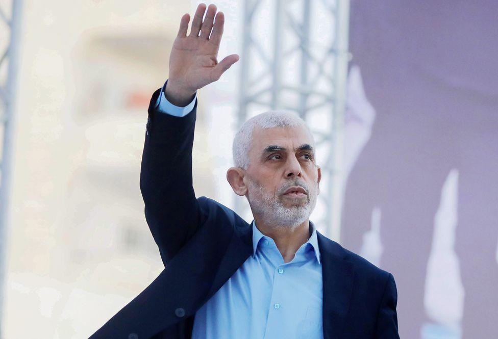 Yahya Sinwar, wearing an open-necked shirt and smart jacket, raising his right hand in the air  