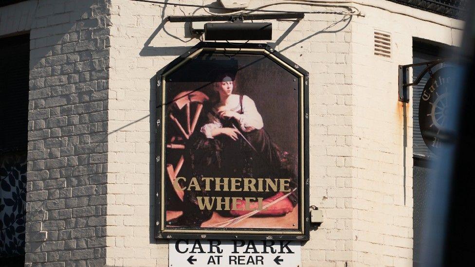 The Catherine Wheel pub in Norwich is a white brick building. The sign on the top of the building is of a woman and in gold lettering it says CATHERINE WHEEL.