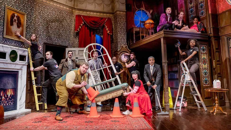 A scene from The Play That Goes Wrong in the Duchess Theatre in London. One actor at the front is holding a traffic comes and a woman in a red dress is lifting up the end of a chaise longue. Other actor on the stage are holding a window frame, climbing a ladder, sweeping the floor, fixing the time on a clock and drilling the set 