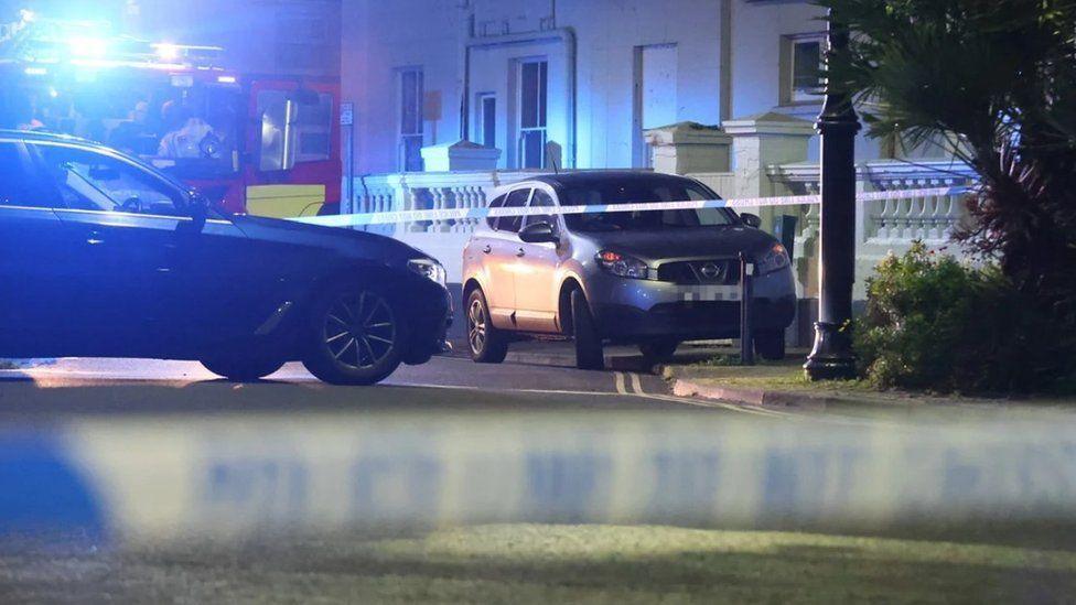 Police tape accross a road with vehicles including a fire engine and a white building in the background - it is dark.