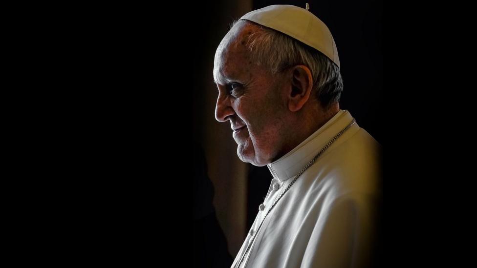 Pope Francis shown in profile looking off to the left, wearing a white cap and robe