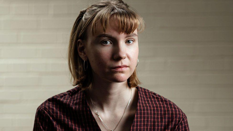 Jenna looks directly at the camera with her head at a very slight angle. The background appears to a plain white painted brick wall. She has light brown hair which is moved behind her ears to show hooped earrings. She wears a necklace and a plaid shirt with no collar and a v-neck.