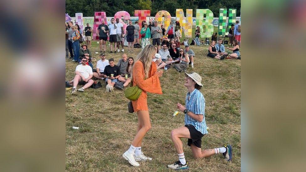 A man down on one knee as he proposes to his girlfriend in front of the Glastonbury sign.