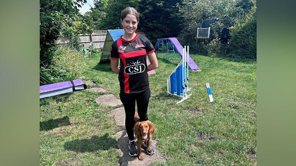 Taylor with dark hair tied down smiling at the camera and wearing a red and black T-shirt and black trousers. A brown cocker spaniel is sitting at her feet. They are in a grassed garden where slides and ramps are arranged in a course.