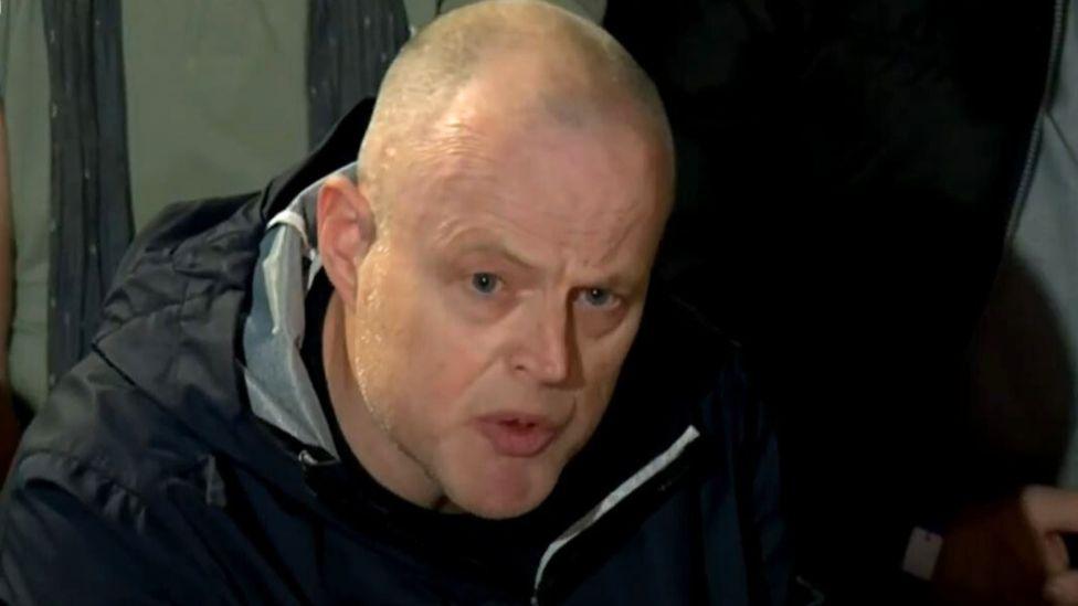 A bald man with a black rain jacket sits hunched forward in a close-up shot 