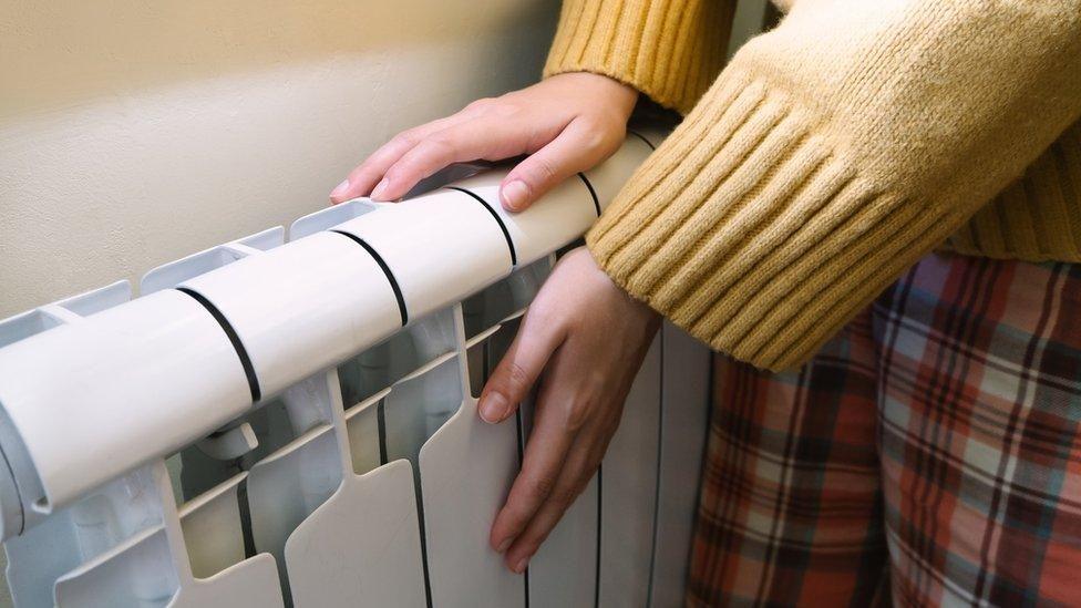 A stock photo of the arms and legs of a person wearing pyjamas and a yellow jumper warming their hands on a radiator. 