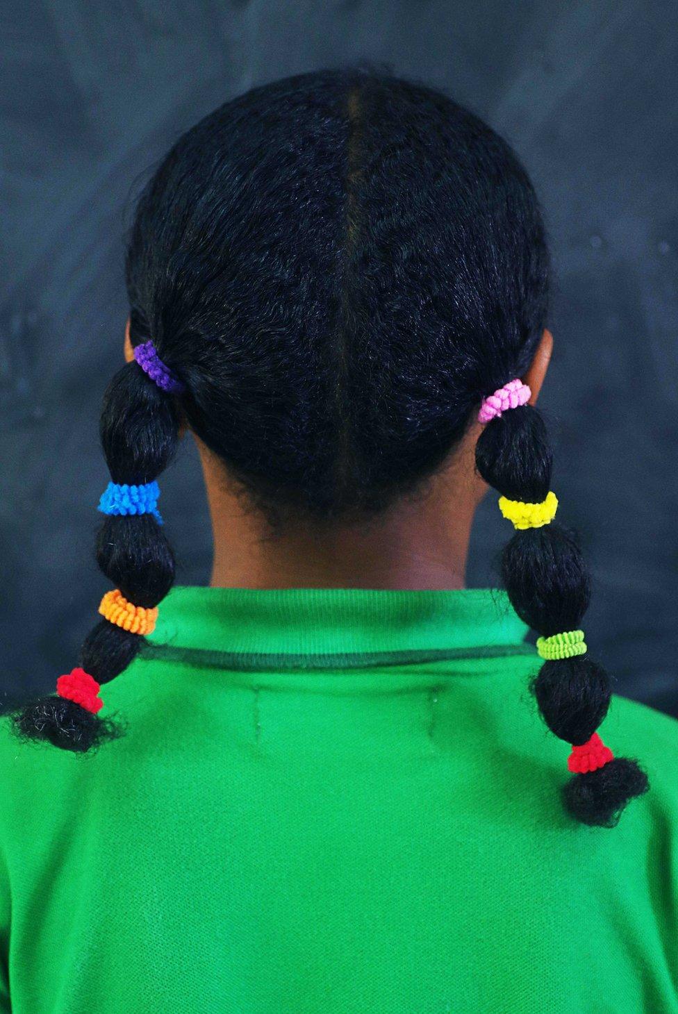 A girl wearing pigtails with several multi-coloured stands with her back to the camera