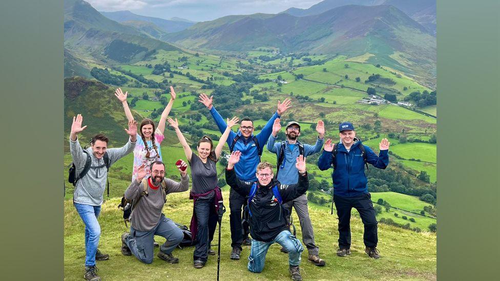 The Stag Walkers posing for a photo on a hill, with mountains in the background. There are nine people with their hands raised in the air, smiling. Two kneel at the front. There are some trees in the valley and a few scattered houses.