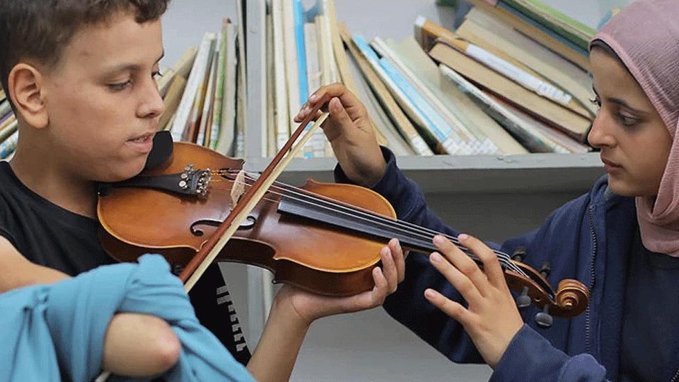 Mohammed Abu Eideh, who lost his right hand in an airstrike, being taught violin by 16-year-old Sama Nijim. Mohammed has a blue scarf tied around his stump to hold the violin bow in place. Sama, in a blue hoodie and pink headscarf, is helping him to position the violin. 