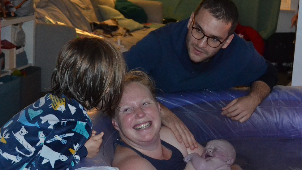 Jocelyn holding her new baby in a birthing pool with her husband and older daughter standing around her