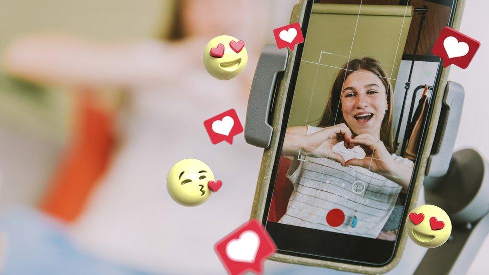 Teenage female influencer showing heart shape while filming through mobile phone with heart emojis surrounding