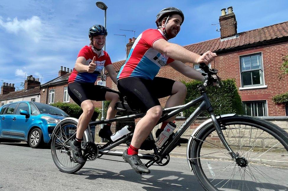 Luke and Elliot riding their tandem bike, giving a thumbs up to the camera