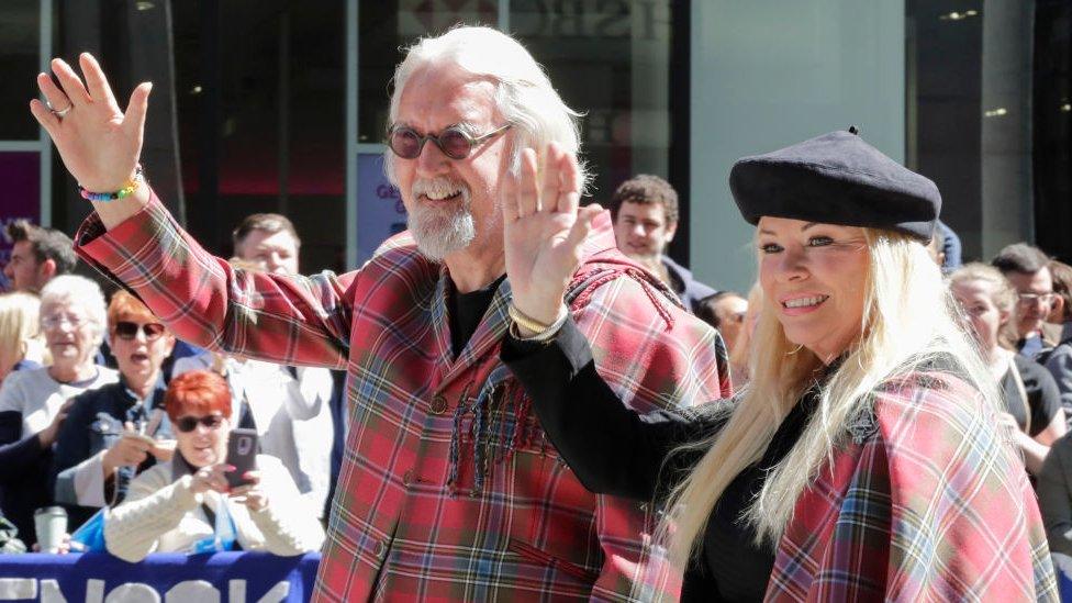 Sir Billy and Stephenson wave in front of a crowd, while wearing matching tartan