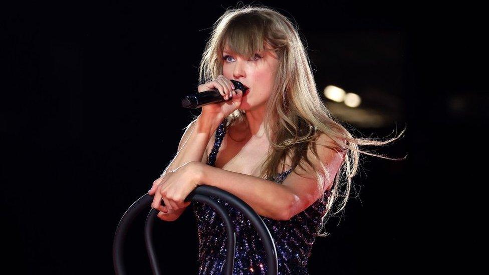 Taylor Swift on her Eras tour, with a micrphone in her right hand hand as she is sat on a black chair, with the back of the chair at the front. She is looking far ahead into the crowd as she sings. She is wearing a dark blue sequined outfit, with a few spotlights visible in the dark background