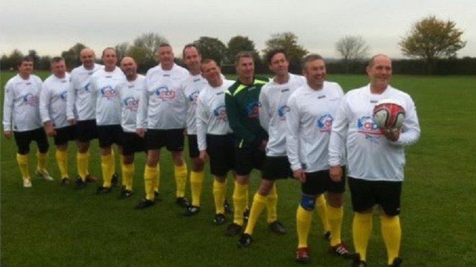 Team of men in their fifties lining up with white shirts, dark shorts and yellow socks. One is holding a ball.