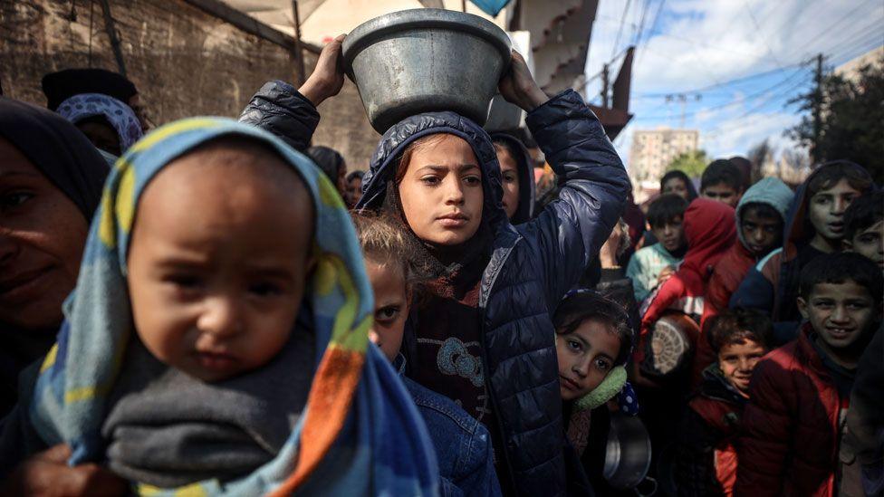 Displaced Palestinian children wait in a line whilst holding bowls as they wait to receive a free hot meal in Deir al-Balah, central Gaza Strip, on 26 November 2024