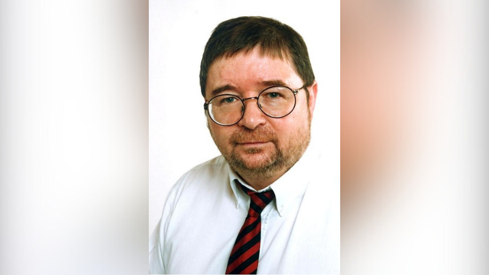 A man wearing glasses and a white shirt with a black and red tie. The backdrop is white.
