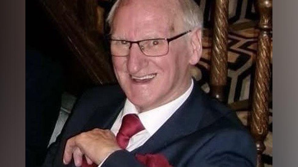 Postmaster Henderson Shaw sitting down smiling and wearing a dark blue suit with a red tie