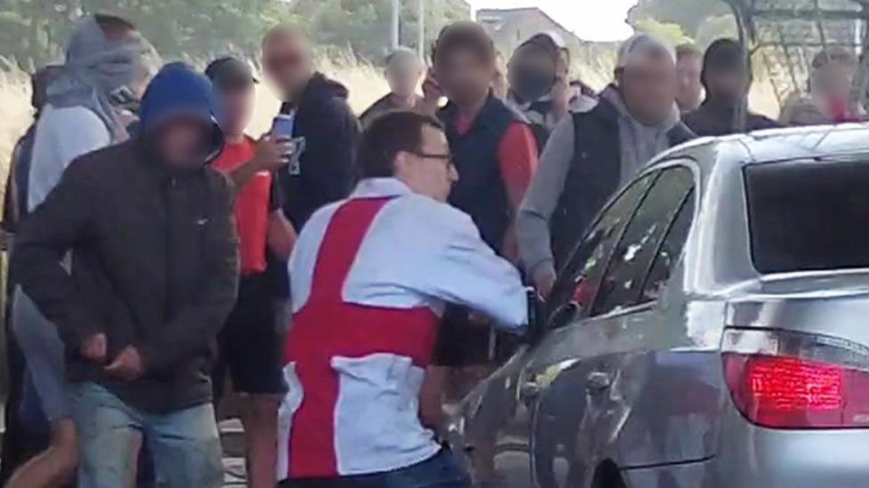 Honey wearing an England flag t-shirt next to the front passenger side of a silver BMW car with a mob in front of the vehicle