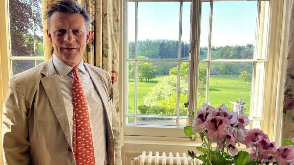 John Coleman wearing a cream suit and red spotted tie stands next to a bunch of pink flowers and in front of a window with views of the countryside in the hall.