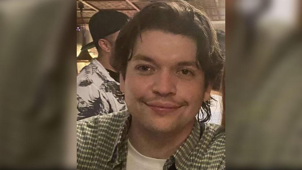 Luis Piovesana wearing a green and white checked shirt over a white t-shirt. He has medium length dark hair and a faint moustache. He appears to be in a bar at night, surrounded by people.