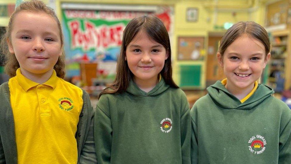 Three school pupils are stood in their school hall.